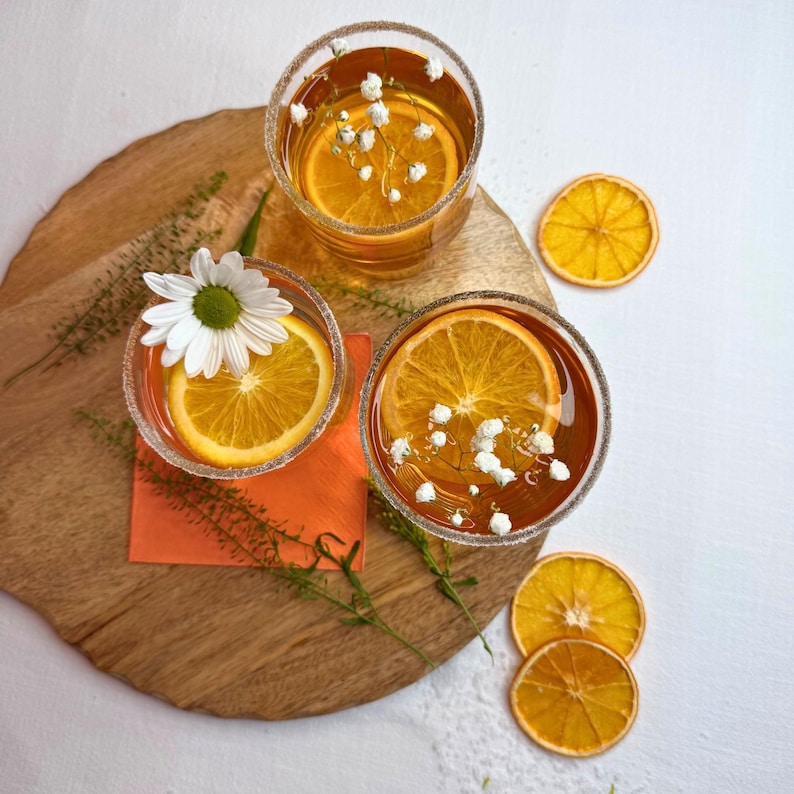 May include: Three clear glasses filled with a golden liquid, garnished with orange slices and small white flowers. The rims of the glasses are sugared. The drinks sit on a wooden board with an orange napkin and dried orange slices.