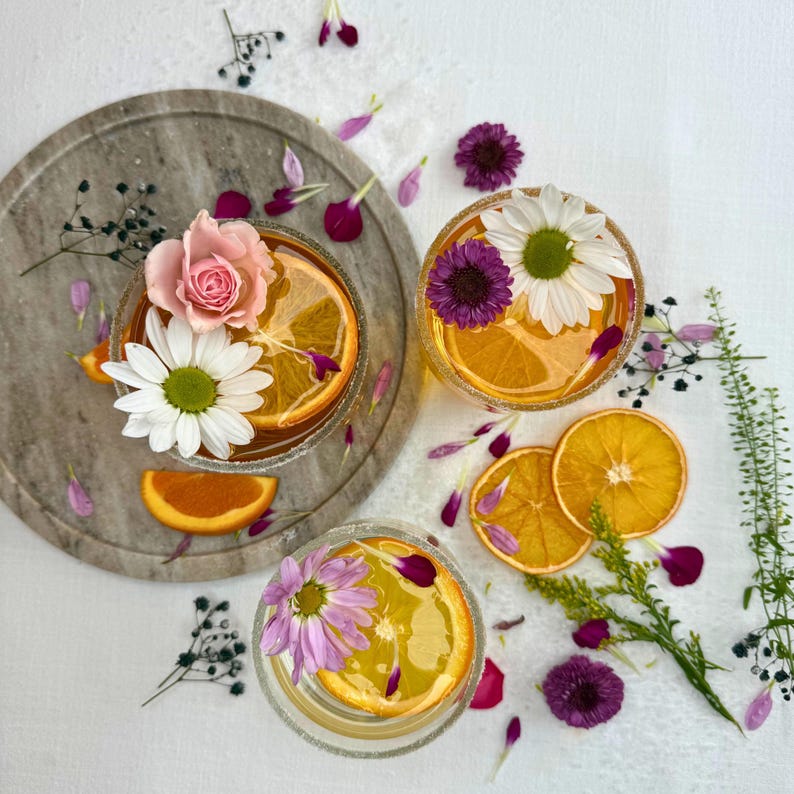 May include: Three clear glasses filled with a golden liquid, garnished with orange slices and colorful flowers. One glass sits on a round, gray marble tray. Scattered around are flower petals and sprigs of greenery, creating a vibrant, refreshing aesthetic.