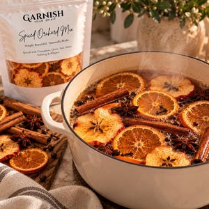 May include: A white pouch labeled Garnish Your Spirit Spiced Orchard Mix containing dried fruit and spices, next to a white pot filled with a steaming liquid, sliced oranges, cinnamon sticks, star anise, and cloves, with loose ingredients nearby.