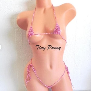 May include: A pink and white crocheted bikini top and bottom. The bikini top is a triangle style and the bottom is a string style. The bikini is very small and revealing. The text "Tiny Pansy" is printed on the model's stomach.