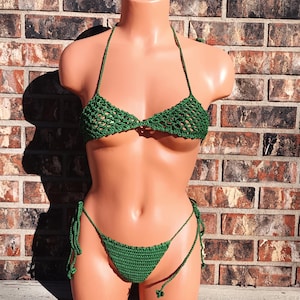 May include: A green crochet bikini set displayed on a mannequin. The bikini top is a triangle style with halter straps, and the bottoms are a string bikini. The set is made of a dark green yarn.
