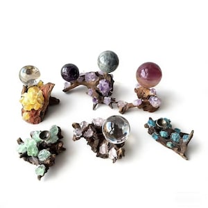 Puede incluir: Una colección de soportes decorativos para esferas de cristal. Cada soporte está hecho de un trozo de madera flotante adornado con cristales coloridos y una esfera de cristal. Las esferas varían en color y tamaño, y la madera flotante tiene un aspecto natural y rústico.