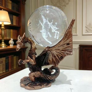 Op de afbeelding: Een bronzen drakenbeeldhouwwerk met een heldere glazen bol. Binnenin de bol bevindt zich een wit, geëtst ontwerp met figuren. De draak heeft gedetailleerde schubben en vleugels.