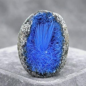 May include: A geode featuring a hollow centre filled with vibrant, needle-like electric blue crystals. The outer shell of the stone is a rough, textured, dark grey and metallic mineral formation that contrasts with the bright blue interior.