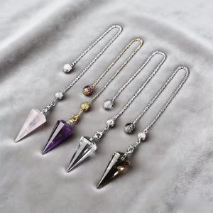 May include: A collection of crystal pendulums with silver and gold chains. The pendulums feature various crystal types, including rose quartz, amethyst, clear quartz, and smoky quartz. Each pendulum has a pointed, conical shape and is suspended from a chain.