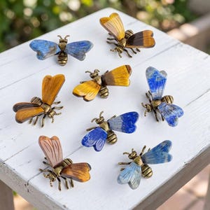 May include: A selection of decorative bee ornaments with wings in blue, yellow, brown, and pink. The bee bodies are bronze-coloured, arranged on a white wooden surface.