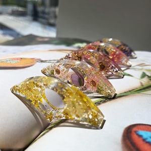 May include: A collection of clear resin rings, each embedded with dried flowers and gold leaf. The rings are arranged in a row, showcasing the variety of floral arrangements and gold accents. The background is a soft, neutral surface.
