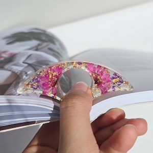 May include: A clear resin book page holder with a curved design. The holder is embedded with pink flowers, gold flakes, and purple accents. The holder is being used to hold open a book.