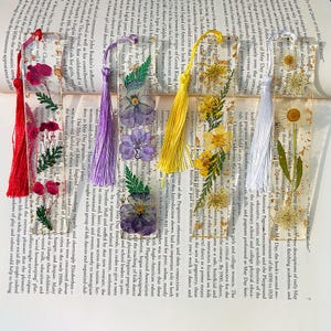May include: A collection of handmade bookmarks with dried flowers and colorful tassels. Each bookmark is clear with pressed flowers and greenery, and a tassel in red, purple, yellow, or white. The bookmarks are on top of an open book.