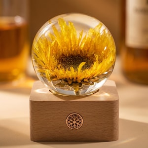 May include: A clear glass sphere containing a yellow sunflower, displayed on a wooden base. The base has a decorative metal emblem. The sunflower is preserved in a transparent material, creating a unique decorative piece.