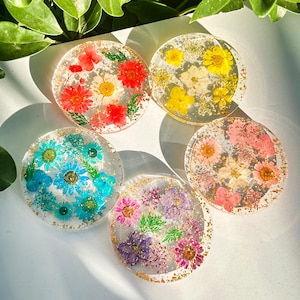 Puede incluir: Cinco posavasos redondos de resina transparente con flores secas de varios colores, incluyendo rojo, amarillo, azul, rosa y morado. Cada posavasos tiene forma circular y está incrustado con copos dorados. Los posavasos están dispuestos sobre una superficie blanca.