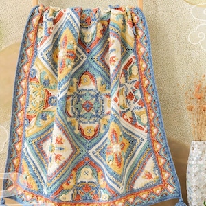 Könnte beinhalten: Eine farbenfrohe, aufwendig gehäkelte Decke mit geometrischem Muster. Die Decke hat Blau-, Gelb-, Orange- und Weißtöne, mit detailliertem Rand und Quasten. Das Design umfasst florale und geometrische Formen.