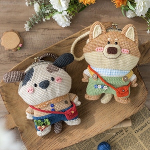May include: Two handmade crochet coin purses shaped like cartoon dogs. One is beige and brown with a black nose and ears, wearing blue overalls. The other is tan and white with a brown nose, wearing green overalls. Both have red crossbody bags.