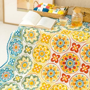 Könnte beinhalten: Eine handgefertigte Häkeldecke mit einem sich wiederholenden Muster aus kreisförmigen Blumenmotiven in Gelb-, Orange-, Blau- und Grüntönen, die mit weißem Garn verbunden sind. Die Decke liegt mit einem offenen Buch und einem Garnkorb aus.