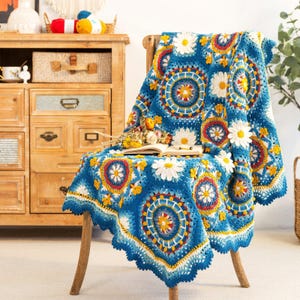 May include: A vibrant blue crochet blanket with a daisy pattern draped over a wooden chair. The blanket features a repeating circular design with white, yellow, and red accents. The edges have a scalloped trim. A book and flowers rest on the blanket.