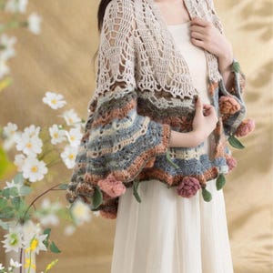 May include: A beige crochet shrug with a floral design on the sleeves. The sleeves feature a mix of blue, brown, and pink colors, with attached pink flower accents and green leaves. The shrug is worn over a white dress.