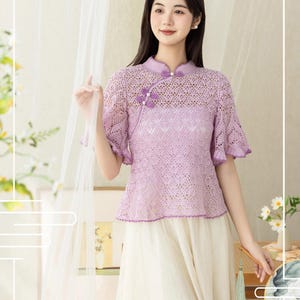 Pode incluir: Uma blusa lavanda de manga curta com um padrão de renda intrincado, gola mandarim e fecho lateral com botões de tecido decorativos. A peça tem uma bainha recortada e é combinada com uma saia longa e fluida na cor creme.
