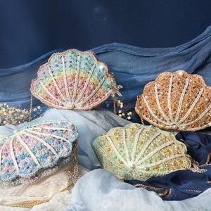 May include: Four seashell-shaped crochet bags in pastel colors. Each bag features a beaded design, a pearl accent, and a gold chain strap. The bags are arranged on a blue and white fabric background, evoking a coastal theme.