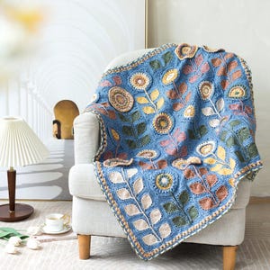 May include: A blue crocheted blanket with a floral design draped over a white armchair. The blanket features colorful flowers and leaves in shades of yellow, pink, green, and brown. The blanket's border is a cream color.