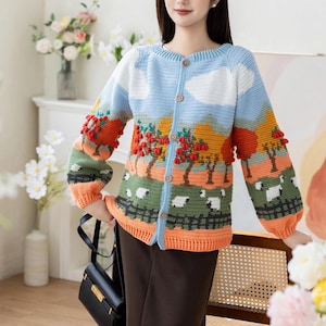 Puede incluir: Cárdigan de crochet azul, blanco, naranja y verde con un diseño de paisaje con árboles, ovejas y nubes. El cárdigan tiene una parte delantera abotonada y mangas largas con detalles de pompón. La modelo lleva pantalones marrones.