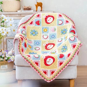May include: A colorful, handmade crochet blanket draped over a chair. The blanket features a patchwork design with squares of yellow, blue, and pink, adorned with strawberry and bunny motifs. The border is trimmed with a scalloped edge in pink and green.