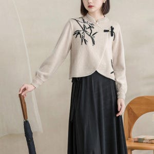 May include: A beige long-sleeved sweater with a wrap-style front, featuring black embroidered bamboo and dragonfly motifs. The sweater has a ribbed collar and cuffs, and is paired with a solid black skirt that has a wrap-around design.