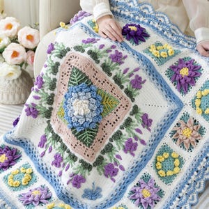 May include: A handcrafted, white crochet blanket with a floral pattern. The blanket has a central square with a blue and white flower, framed by pink and green details. The border is blue with a scalloped edge, and it's decorated with purple and yellow flowers.