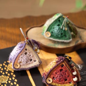 May include: Three triangular crochet pouches featuring intricate openwork patterns. One pouch is purple and beige, one is dark green, and one is a gradient of red, orange, and yellow. Each pouch includes a small wooden button and a decorative tassel.