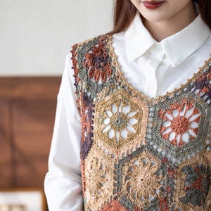 May include: A sleeveless crochet vest featuring a pattern of interconnected hexagonal motifs in shades of rust, sage green, beige, and dark brown, worn over a crisp white collared button-down shirt with a pointed collar and visible front buttons.