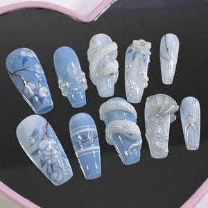 Puede incluir: Una colección de diez uñas artificiales con varios diseños. Las uñas son principalmente de color azul claro y blanco, con diseños florales, detalles de perlas y detalles plateados. Algunas uñas presentan un diseño de serpiente.