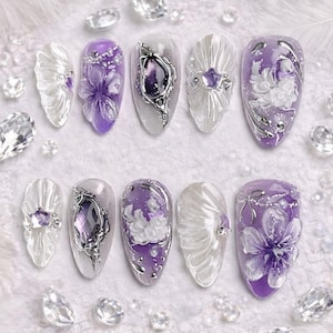 May include: A collection of artificial nails with detailed designs. The nails showcase a range of colours, including white and purple, with embellishments such as rhinestones, floral patterns, and metallic accents. The nails are arranged on a white surface with scattered crystals.