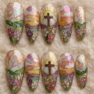 Könnte beinhalten: Ein Set aus zehn langen, oval geformten Kunstnägeln. Jeder Nagel zeigt ein Buntglasdesign mit einem Kreuz, Sonnenuntergang und floralen Elementen in Rosa, Lila, Grün und Gold. Die Nägel haben ein glänzendes Finish und sind vor einem weichen, weißen Hintergrund platziert.