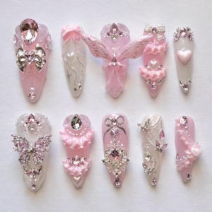 May include: A collection of ten artificial nails, each uniquely designed with pink, white, and silver embellishments. Features include bows, hearts, butterflies, and rhinestones. The nails are oval-shaped and showcase intricate, decorative nail art.