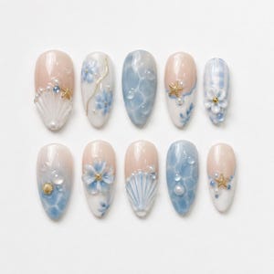 May include: A set of ten almond-shaped press-on nails featuring a coastal theme with light blue and nude gradients. The designs include 3D white seashells, gold starfish charms, small white pearls, blue floral accents, and clear water droplet effects.