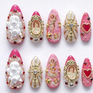 May include: Ten artificial nails in a variety of designs. The nails are oval-shaped and feature pink and white bases. Decorations include cherubs, framed portraits, gold crosses, and heart shapes, embellished with red gems and pearls.