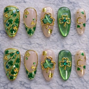 May include: A set of ten artificial nails with a St. Patrick's Day theme. The nails feature green, gold, and nude colors, with clover and gem embellishments. Some nails have a gold outline, while others have a green base.