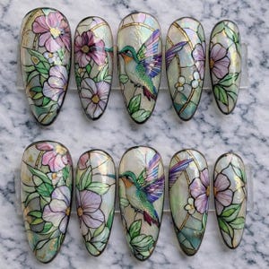 May include: Set of ten oval-shaped false nails with a stained-glass effect. Each nail features a design of flowers, leaves, and a hummingbird. The color palette includes pink, purple, green, and gold accents on a pearlescent background.