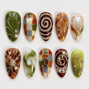 May include: A collection of ten artificial nails with various designs. The nails feature a range of colors, including green, brown, red, and gold, with intricate patterns, gems, and pearl accents. Each nail showcases a unique artistic design.