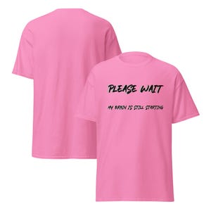 May include: A bright pink short-sleeved t-shirt featuring the black text "PLEASE WAIT" on the top line and "MY BRAIN IS STILL STARTING" on the second line. The shirt is displayed from both the front and back angles, showing a plain back.