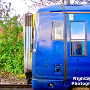 May include: A vibrant blue train carriage with a yellow stripe, featuring windows and a door. The background shows green foliage, a building, and railway tracks. The image is labelled "NightSpark Photography".