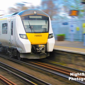 May include: A high-speed passenger train in motion, with a white, yellow, and blue livery. The front of the train displays the number 700025 on a yellow panel. The image is blurred, indicating speed, and the train is on railway tracks.