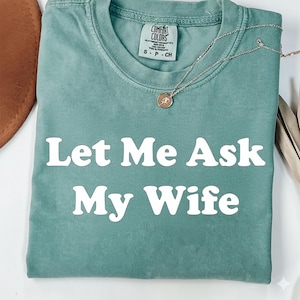 May include: A teal green t-shirt with the text "Let Me Ask My Wife" in white block letters. A silver necklace with a small pendant rests on the shirt. The shirt has a Comfort Colors tag.