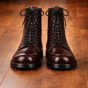 May include: A pair of burgundy leather boots with black laces. The boots have a classic design with a rounded toe and a sturdy black sole. The boots are displayed on a wooden surface.