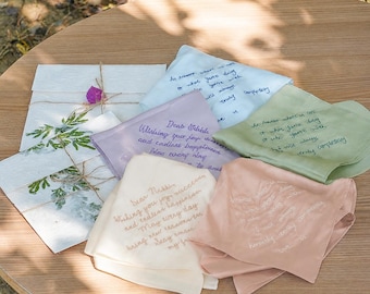 Könnte beinhalten: Eine Sammlung von fünf quadratischen Stofftaschentüchern in Pastelltönen wie Hellblau, Lavendel, Salbeigrün, Creme und Altrosa, jeweils mit gesticktem kursivem Text. Ein weißer Papierumschlag, verziert mit grünen Blättern und Bindfaden, liegt bei.