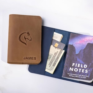 May include: A brown leather wallet with a horse head design and the name "JAMES" embossed on the front. Next to it, an open blue wallet holds cash, a credit card, and a "FIELD NOTES" memo book with a landscape image.
