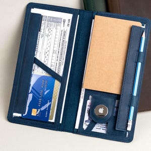 May include: Open navy blue leather folio with a notepad, credit card, and documents. A pencil is secured in a leather loop. Two additional leather folios in green and brown are visible in the background. The folio is designed to hold various items.