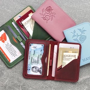 May include: An assortment of leather wallets in various colors, including green, pink, blue, and burgundy. Some wallets are open, displaying cards and identification. The wallets feature embossed floral designs and the name "MAYA" on one.