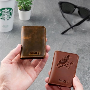 May include: Two brown leather wallets are held in front of a gray background. One wallet is embossed with the name "JAMES", the other with a raven design and the name "MIKE". A Starbucks cup, sunglasses, and a small plant are in the background.