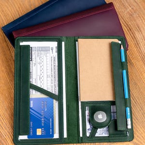 May include: A green leather folio with a credit card, notepad, and pencil. The folio is open, revealing interior pockets and a small Apple AirTag. Two additional folios in navy and burgundy are stacked behind it on a wooden surface.