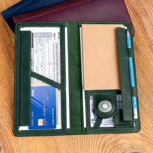 May include: A green leather folio with a credit card, notepad, and pencil. The folio is open, revealing interior pockets and a small Apple AirTag. Two additional folios in navy and burgundy are stacked behind it on a wooden surface.